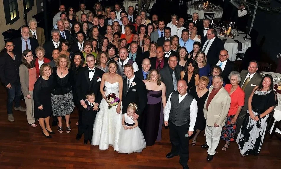 Large group of people gathered at a wedding reception, with the bride and groom in formal attire at the center.