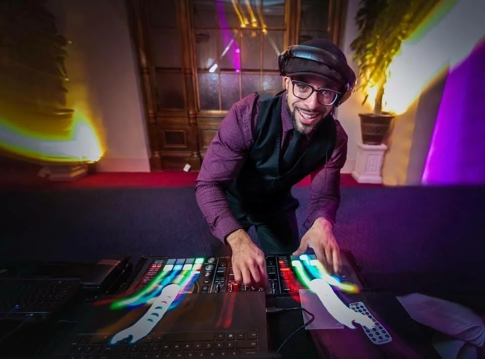 A DJ wearing glasses, a black vest, and a purple shirt is at a mixing console, smiling at the camera in a decorated room with colorful lights.