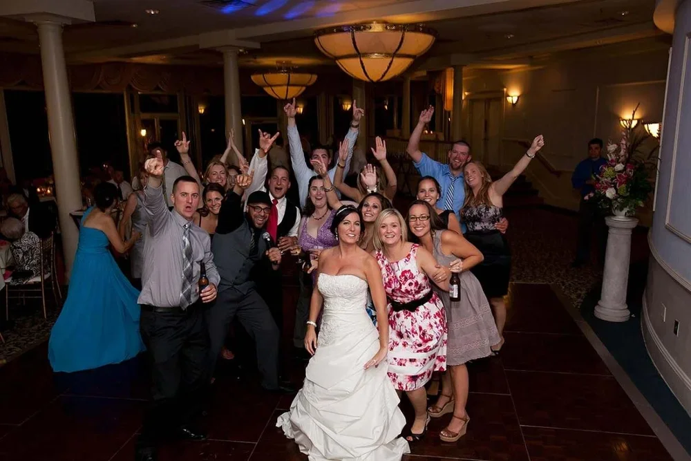 Group of people celebrating at a wedding reception, with the bride in a white wedding dress at the front center, all smiling and raising their hands in celebration.