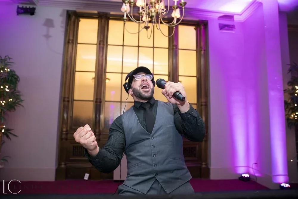 A man singing passionately into a microphone at an indoor event, wearing a grey vest and black shirt, with headphones, in front of a large window with purple lighting.