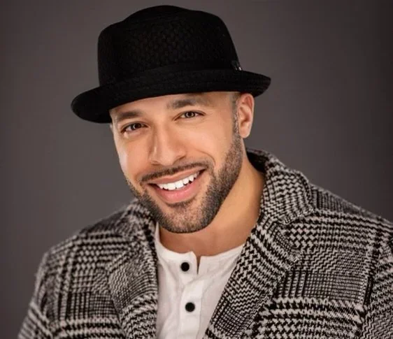 A smiling man wearing a black hat, a checkered blazer, and a white shirt with black buttons, against a gray background.