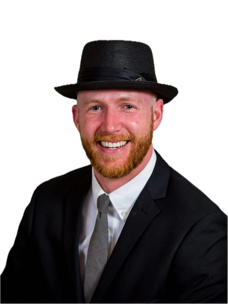 A man with a red beard wearing a black hat, black suit, white shirt, and gray tie, smiling against a black background.
