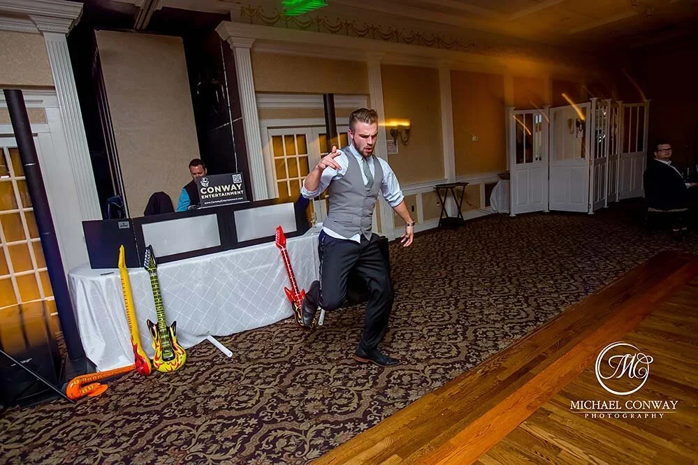 A man in a vest and tie dancing on a carpeted ballroom floor at an indoor event with a DJ booth in the background, decorated with inflatable guitars.