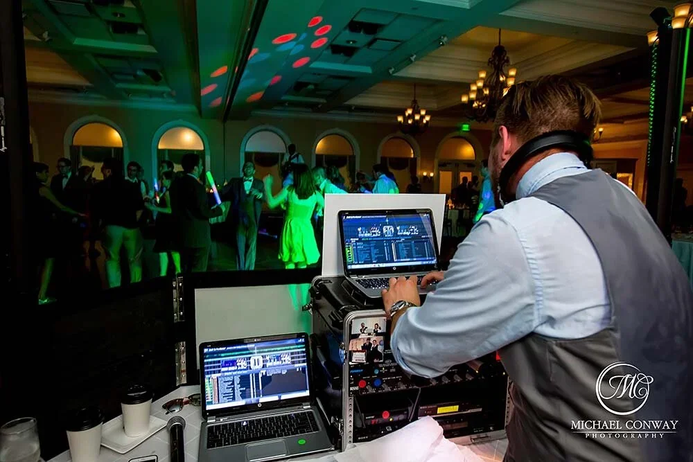DJ wearing headphones and operating mixing equipment at a dance party, with people dancing and colorful lighting in the background.
