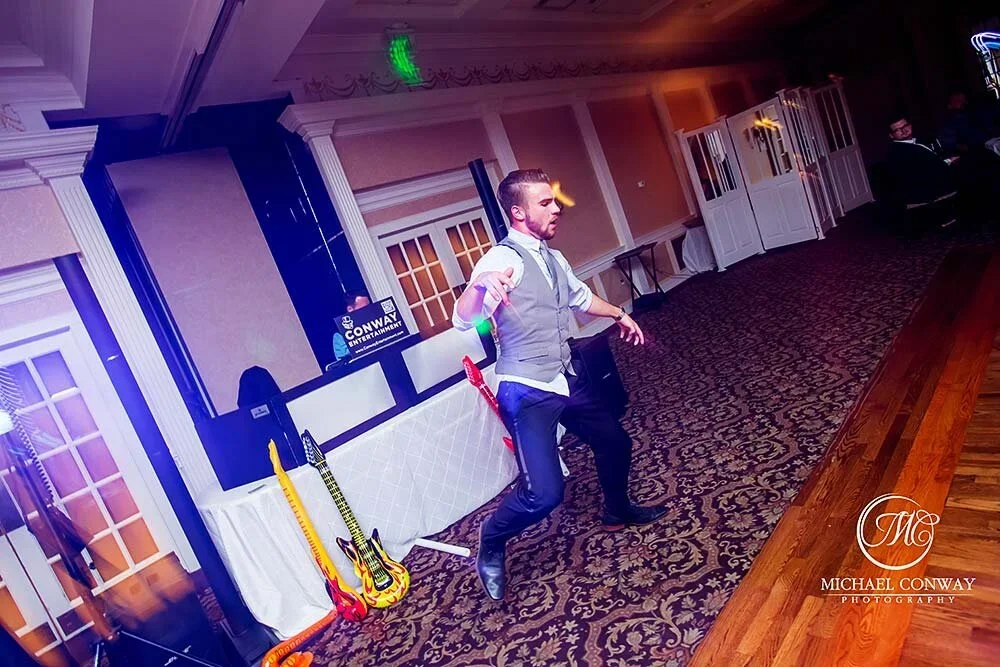 A man is dancing at a wedding reception or party, with a DJ booth, guitars, and decorative elements in the background.