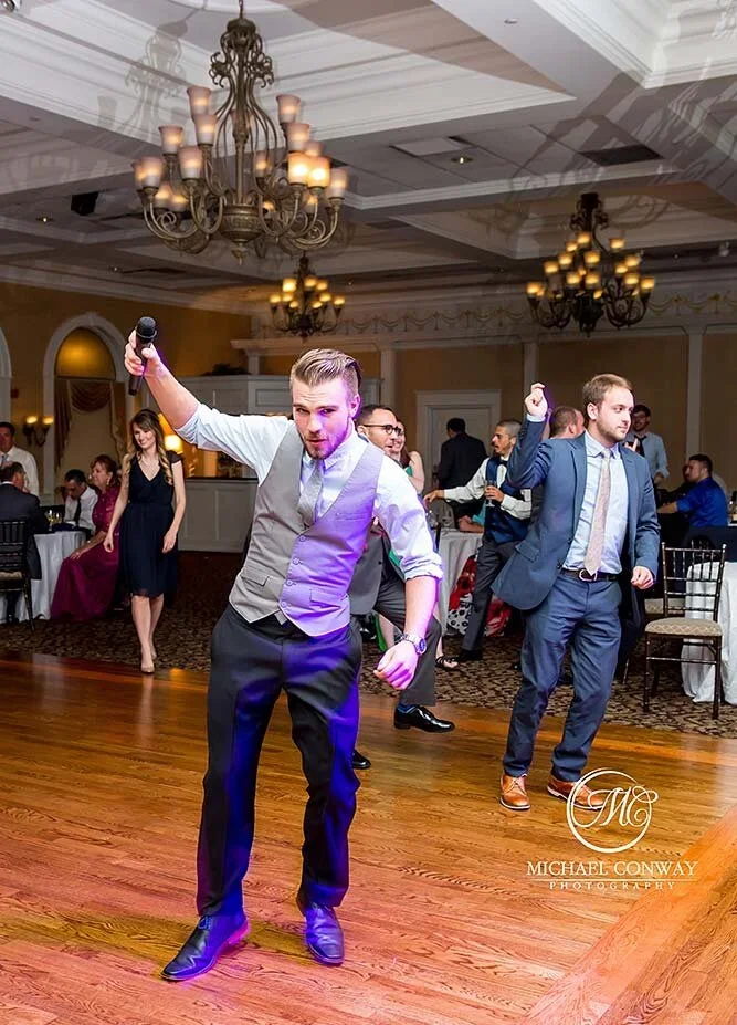 People dancing and celebrating in a formal venue with chandeliers, some seated at tables, with a young man in a vest and dress shirt holding a microphone and dancing.