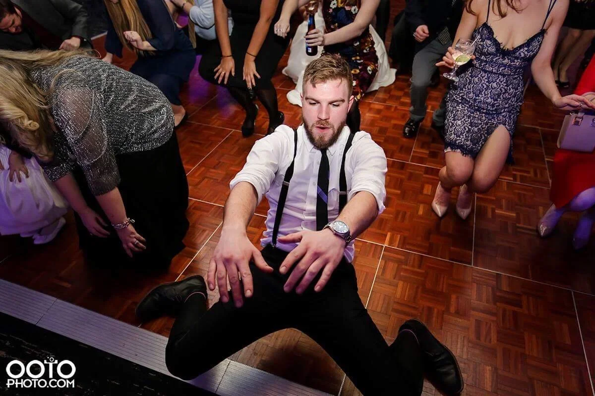 A man with a beard and short blond hair wearing a white shirt, black suspenders, and a black tie dancing on a wooden dance floor at a party or wedding, surrounded by women in dresses and women in formal attire.
