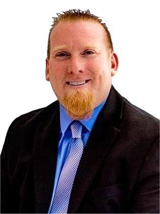 A man with a red beard and mustache, wearing a dark suit jacket, blue dress shirt, and tie, smiling at the camera against a plain background.