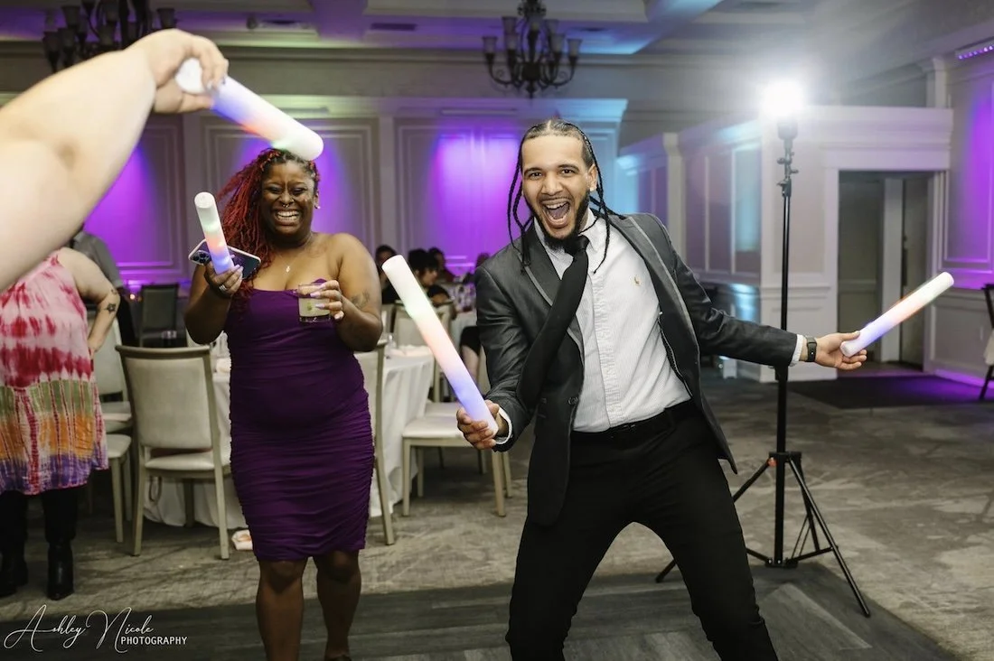 People dancing and having fun at a party with colorful lighting, some holding glow sticks, in a decorated banquet hall.
