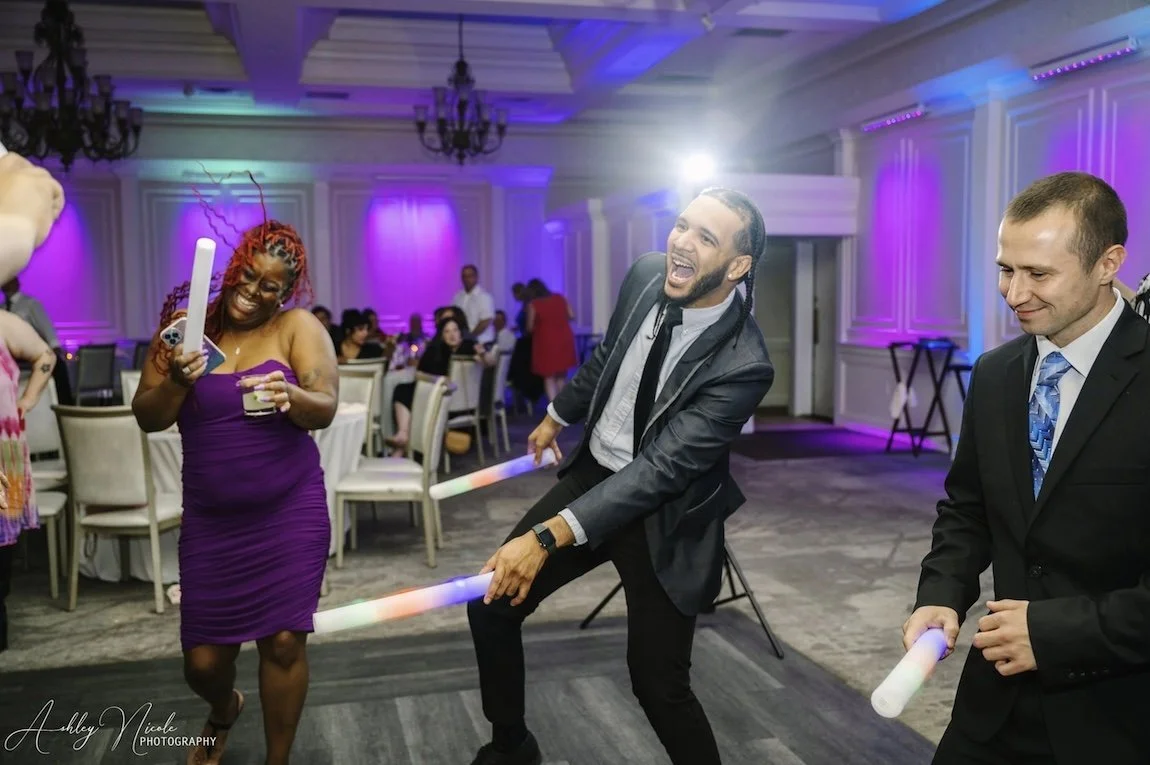 People dancing and enjoying a celebration with colorful lights, including women and men holding glow sticks, in a decorated event hall.