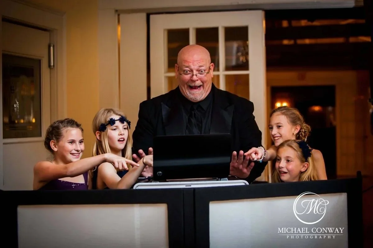A man in a black suit and glasses on a gaming computer with four young girls around him, all smiling and showing excitement.