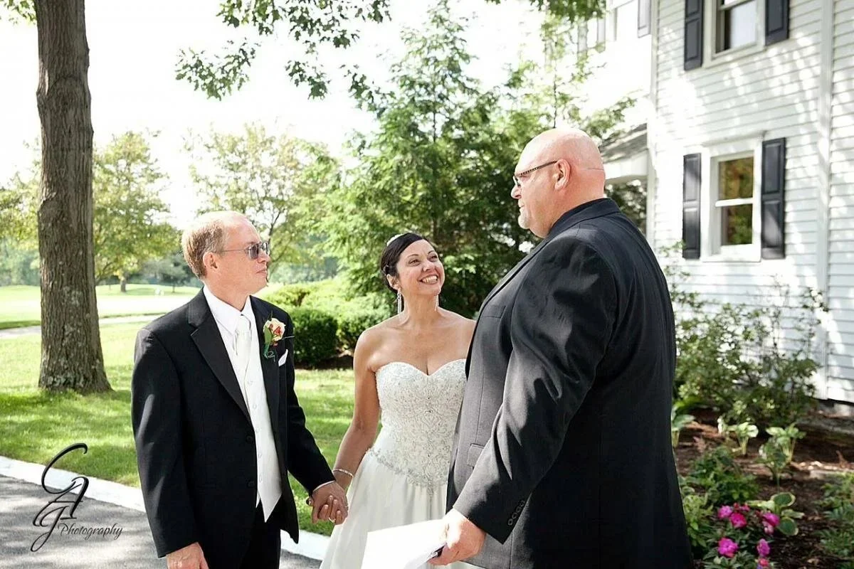 A bride and groom holding hands in front of an officiant during a wedding ceremony outdoors on a sunny day, with trees and a white building in the background.