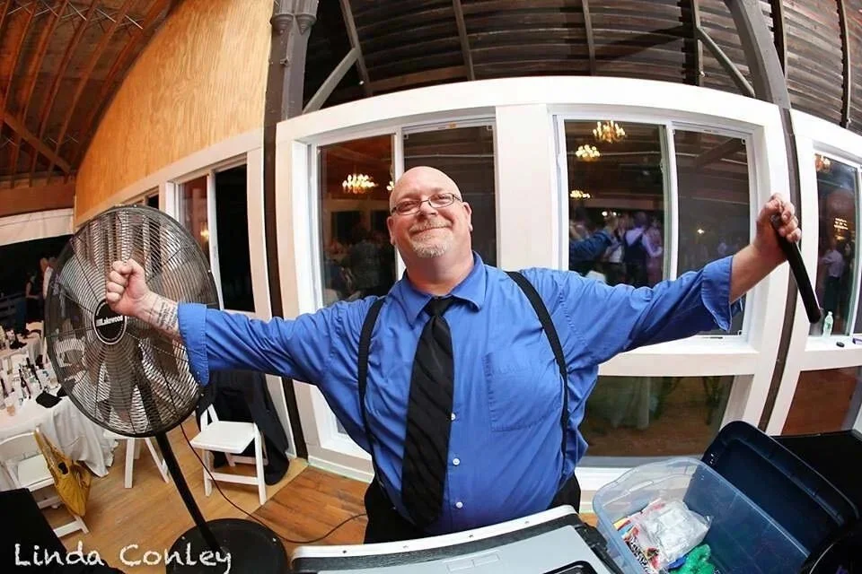 A man wearing a blue shirt, black tie, and suspenders stands with arms outstretched, smiling, in front of a windowed building at an indoor event. He holds a fan in one hand and a tool in the other, with a table and a plastic container filled with mis