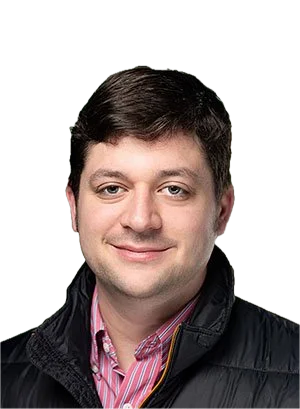 A man with short dark hair, wearing a black jacket over a pink and white striped shirt, smiling at the camera.
