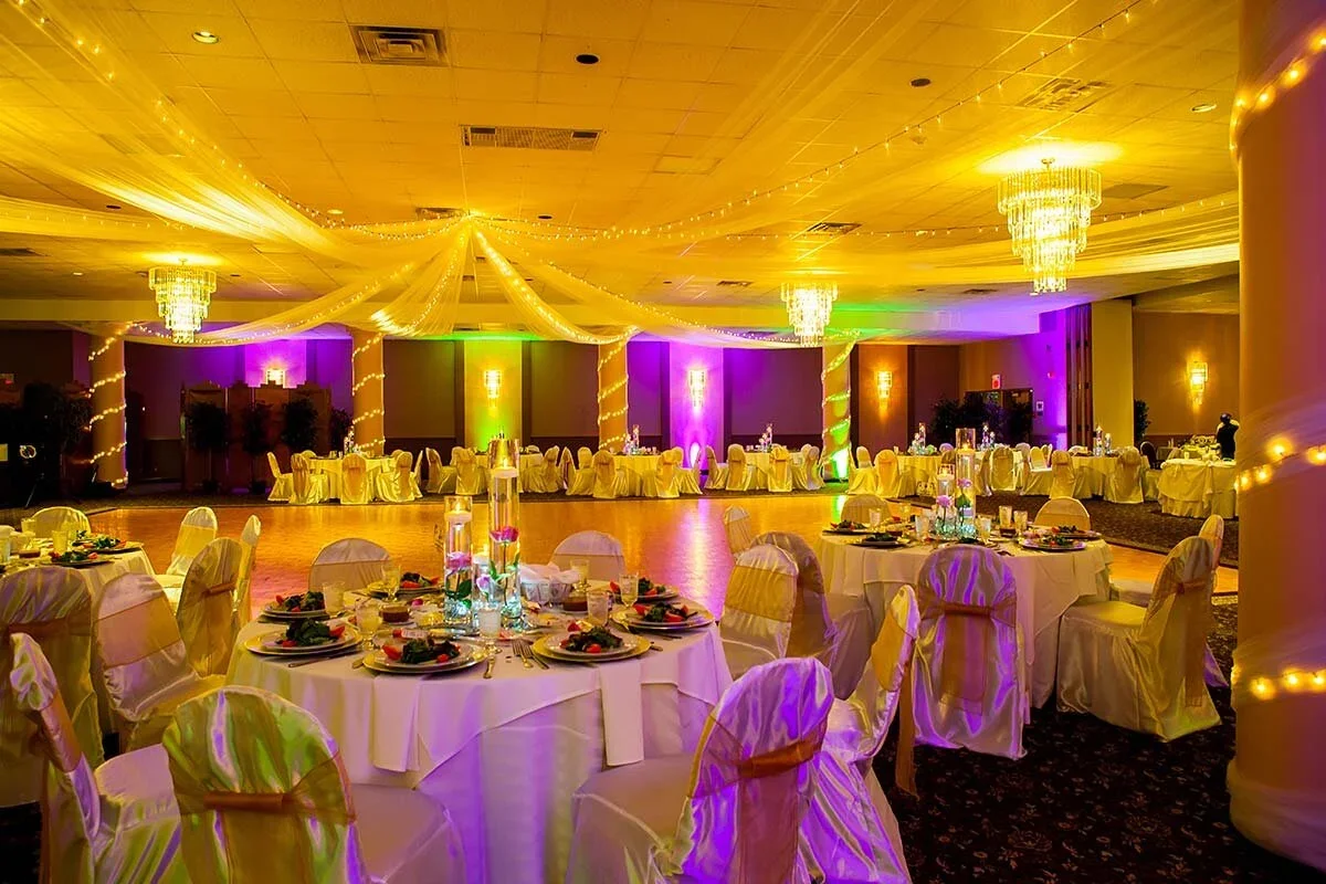 A decorated banquet hall with round tables covered in white tablecloths, chairs with white covers and bows, and a dance floor. The ceiling is adorned with draped fabric and fairy lights, with multiple chandeliers hanging down. The walls are lit with colorful uplighting greens, purples, and warm yellow light.