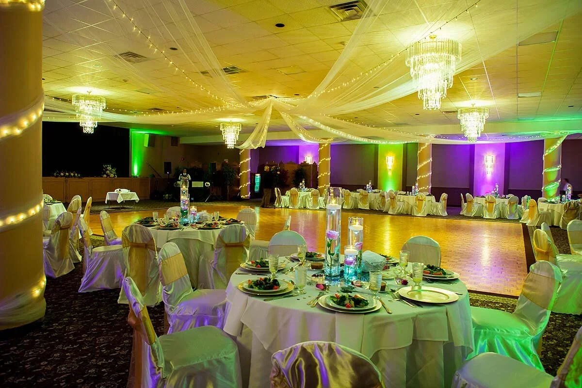 Elegant event hall decorated with white drapes, string lights, and chandeliers, featuring round tables with white tablecloths, gold chairs, and centerpieces with candles and flowers, set for a banquet or wedding reception.