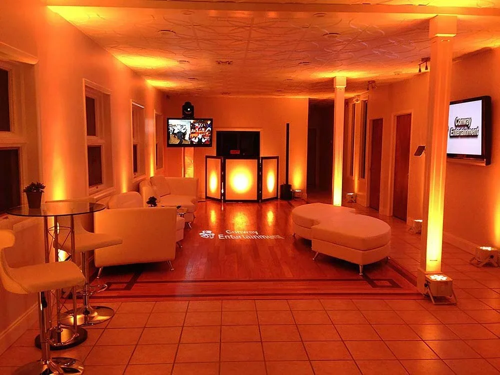 A cozy, dimly lit event lounge with white seating, high-top tables, a DJ booth with orange lighting, and wall-mounted TVs, creating an inviting atmosphere for entertainment.