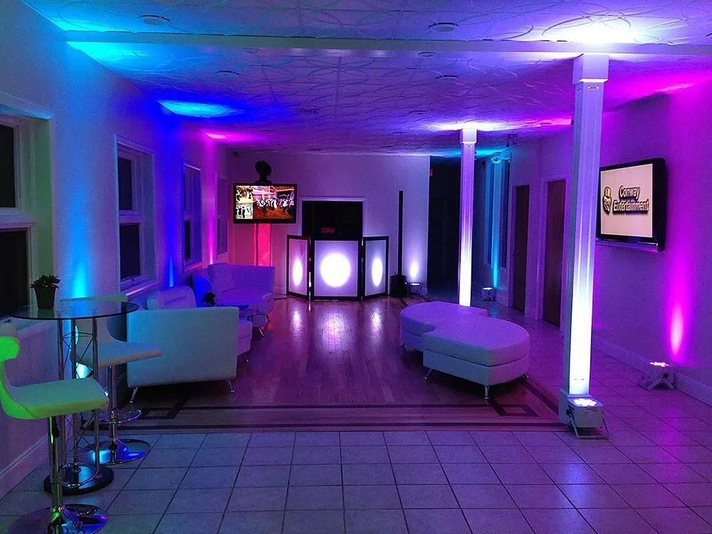 An empty indoor event space with colorful LED lighting, white furniture, a DJ booth, and a TV screen on the wall.