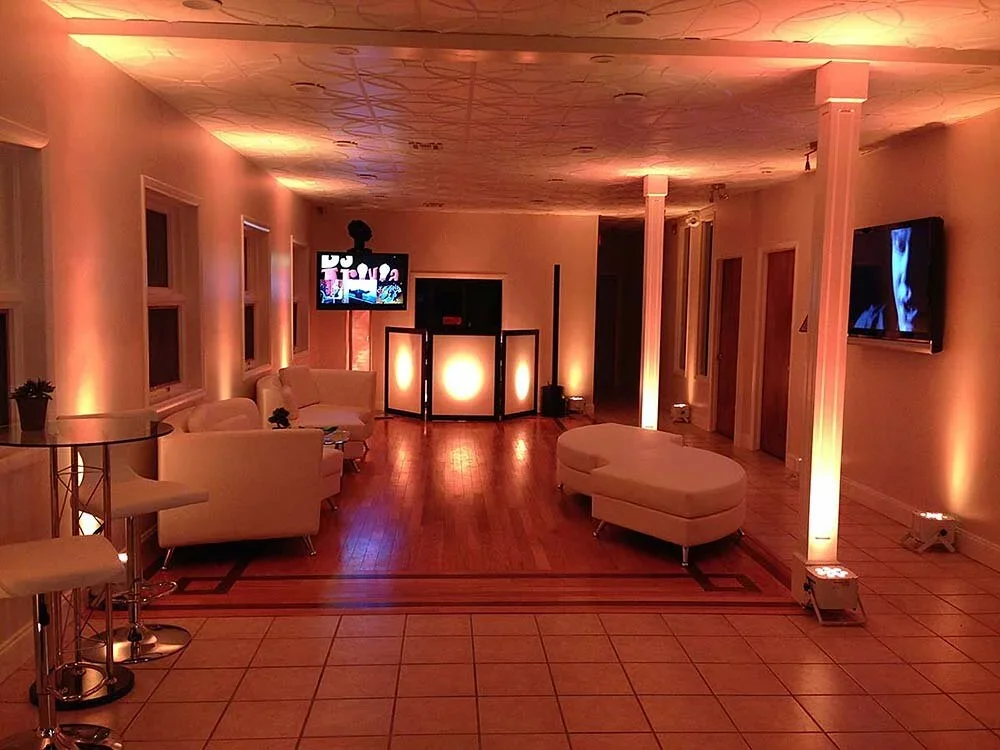 A dimly lit lounge area with white couches, a curved ottoman, a TV mounted on the wall, and a DJ booth in the background, illuminated with soft orange and blue lighting.