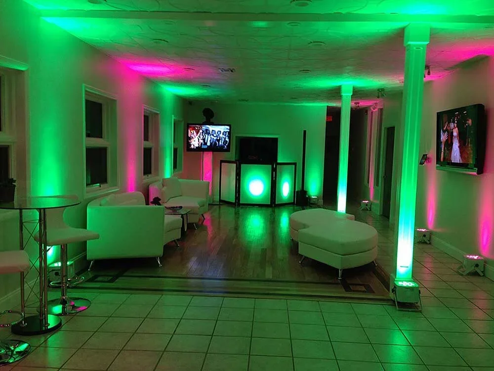 A party lounge with colorful green and pink lighting, white couches, and two flat-screen TVs on the walls.