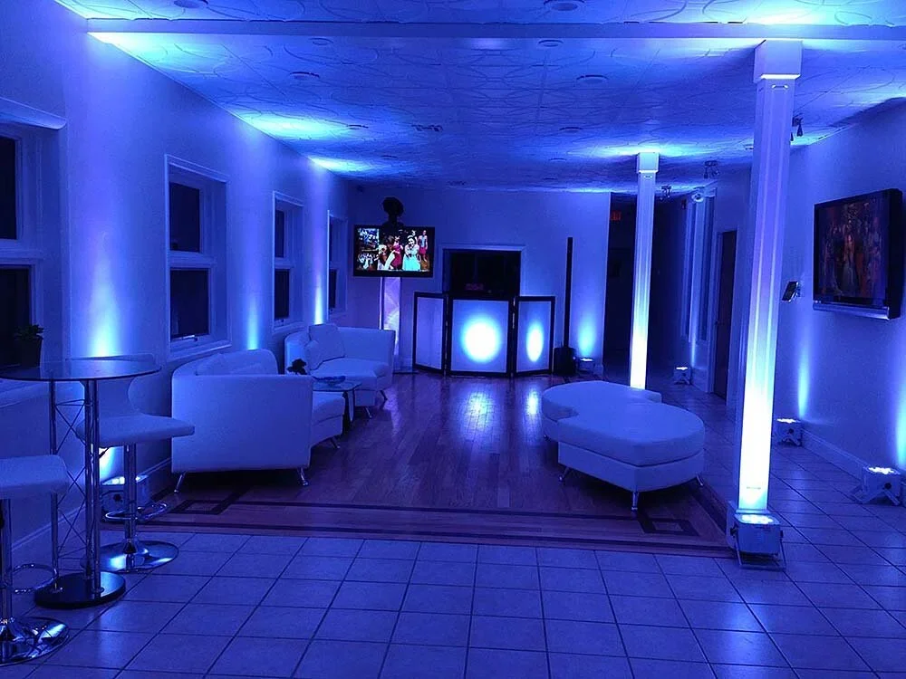 A dimly lit room with blue lighting, white furniture, a DJ booth, a flat-screen TV, and several narrow windows, suggesting a lounge or event space.