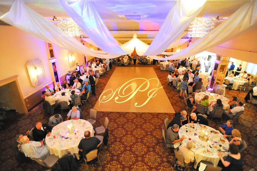 Wedding reception with guests seated at round tables and a large dance floor in the center with monogram initials 'JPG' projected on the floor, decorated with draped white fabric and chandeliers.