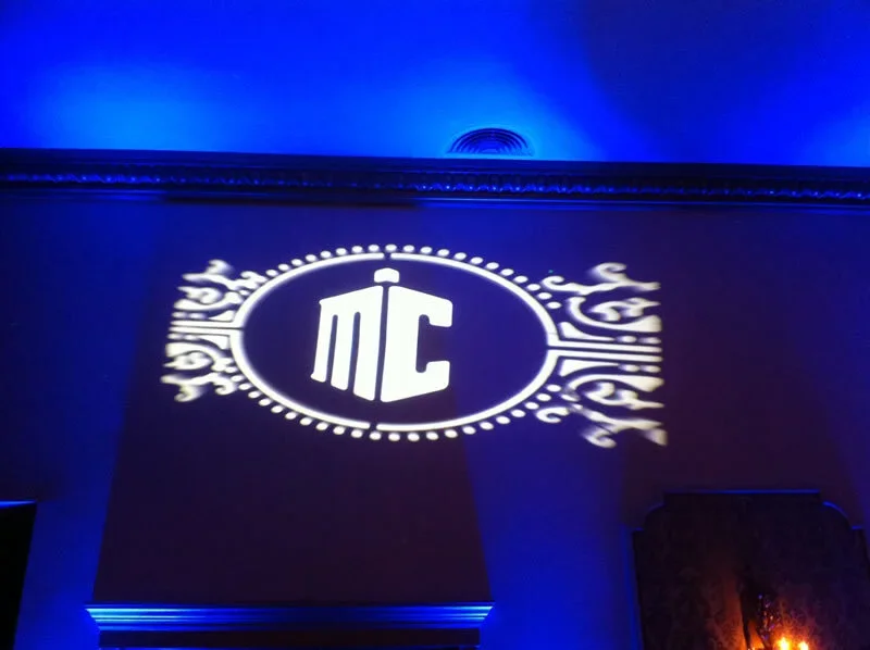 Projection of a logo with stylized initials 'MC' and decorative elements on a wall with blue lighting.