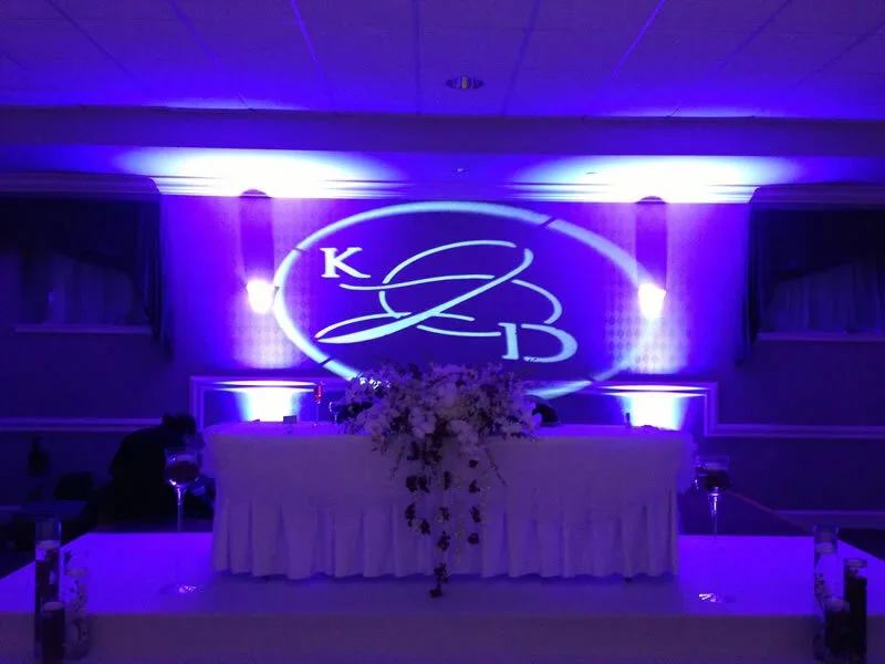 Wedding reception with illuminated monogram 'K & B' projected on the wall, decorated with flowers and purple lighting.