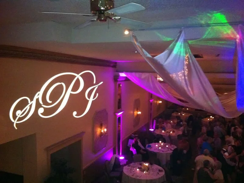 Elegant dimly lit banquet hall with tables, decorative drapery, and purple lighting. A monogram on the wall displays the initials 'S.P.G.'.