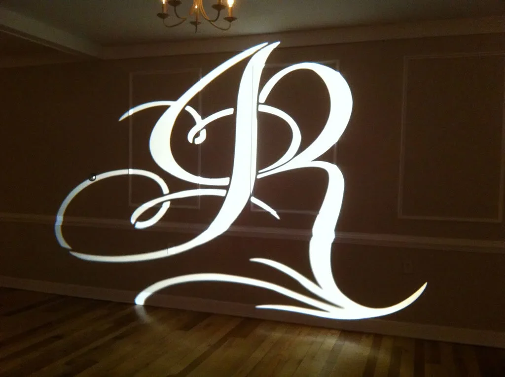 Illuminated white monogrammed letter B with decorative swirls projected on a wall in a dimly lit room.
