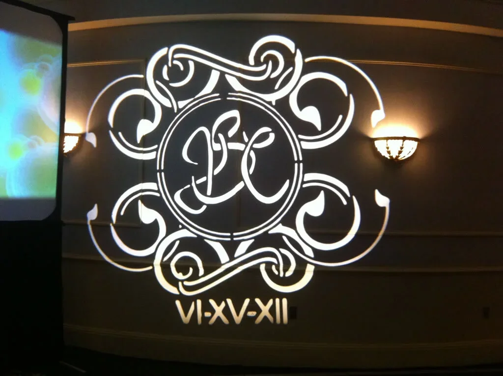 Decorative wall with monogram initials and Roman numerals VI-XV-XII, featuring an ornate design and hanging wall lights.
