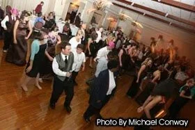 People dancing at a social event in a large hall with wooden floors, some dressed in formal attire.