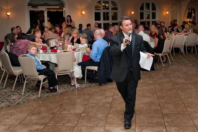 A man in a tuxedo speaking into a microphone at a formal event with many seated guests dining in the background.