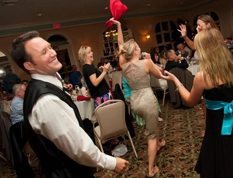 People dancing and celebrating in a banquet hall, some clapping and smiling, with a woman holding a red cloth or floatie in the air.