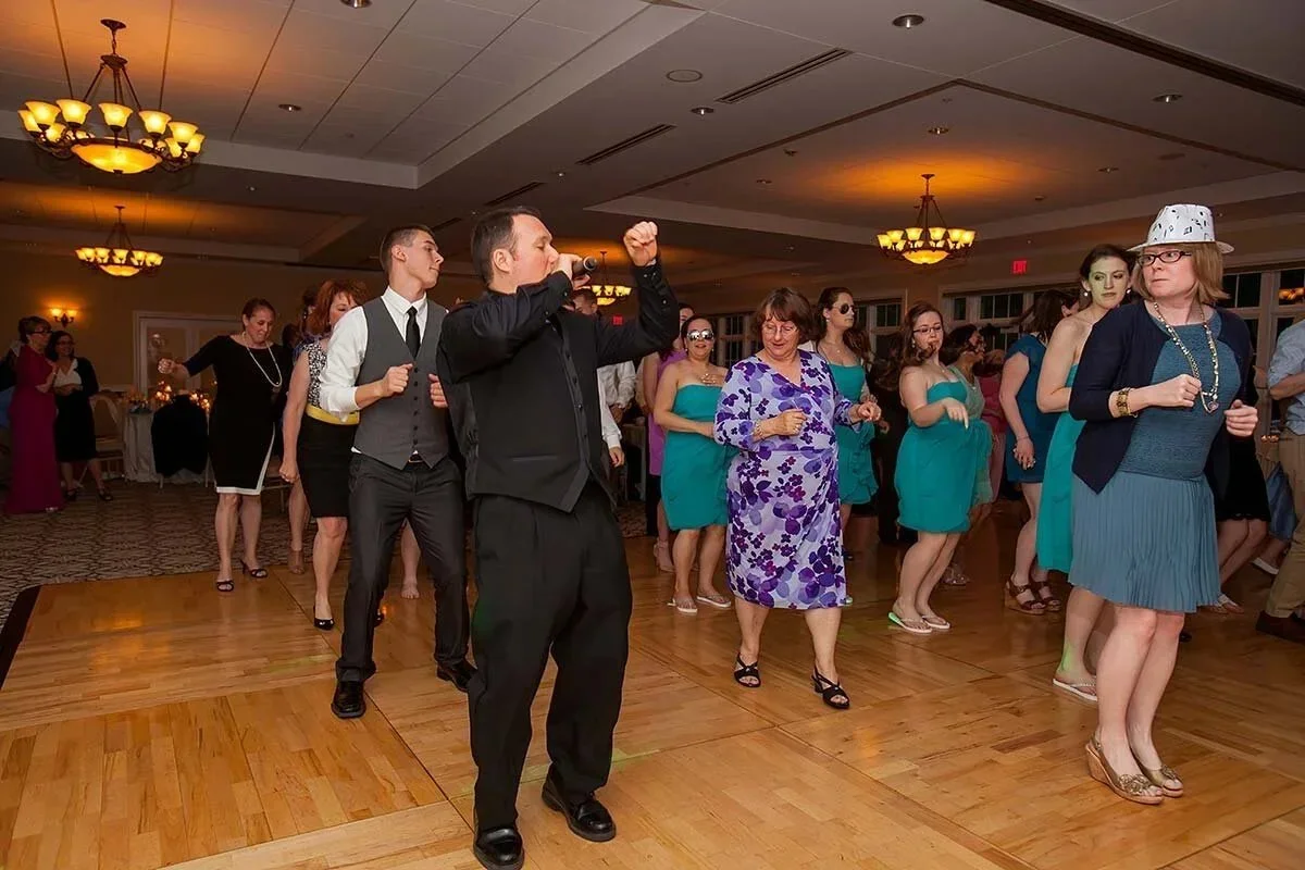 People dancing on a wooden dance floor in a banquet hall, with a man in a black shirt singing into a microphone at the front, surrounded by women in dresses, some wearing glasses and hats.