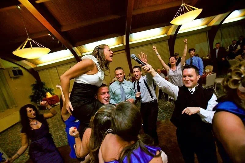 People celebrating at a party, with a woman being lifted in the air, singing into a microphone, and others cheering and dancing around her in a banquet hall.