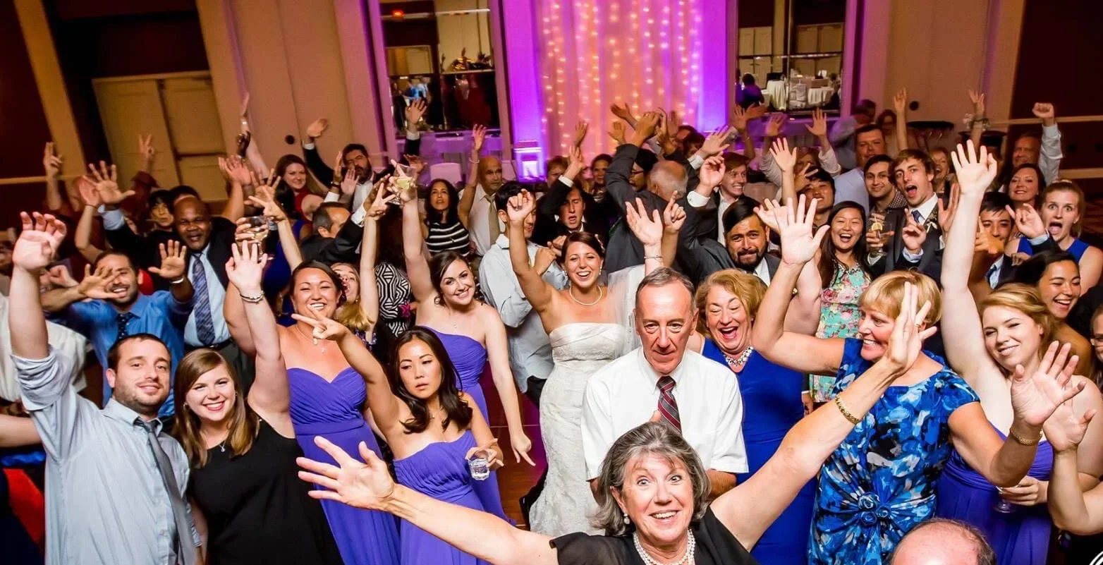 A large group of people celebrating at a party or wedding reception, with many smiling, raising their hands, and holding drinks in a decorated hall with purple lighting.