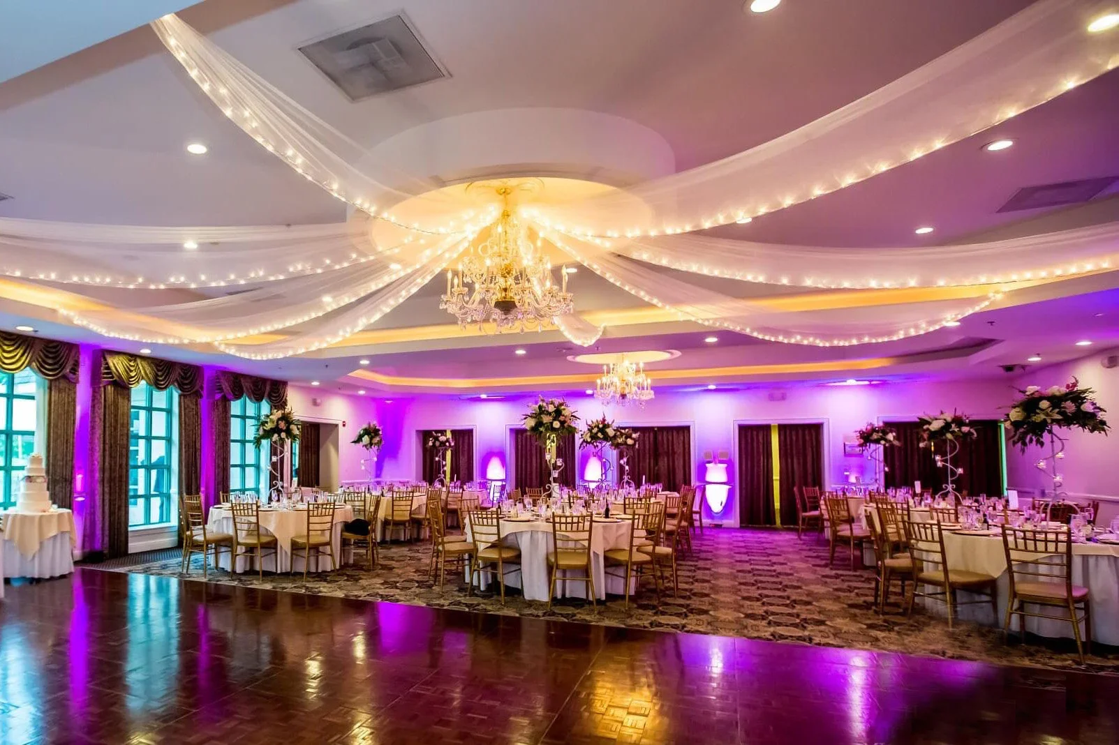 Elegantly decorated banquet hall with round tables covered in white tablecloths, tall floral centerpieces, and gold chairs. The ceiling is draped with white fabric and string lights, and a chandelier hangs in the center. The room features purple lighting, large windows with curtains, and a wedding cake on a small table to the side.