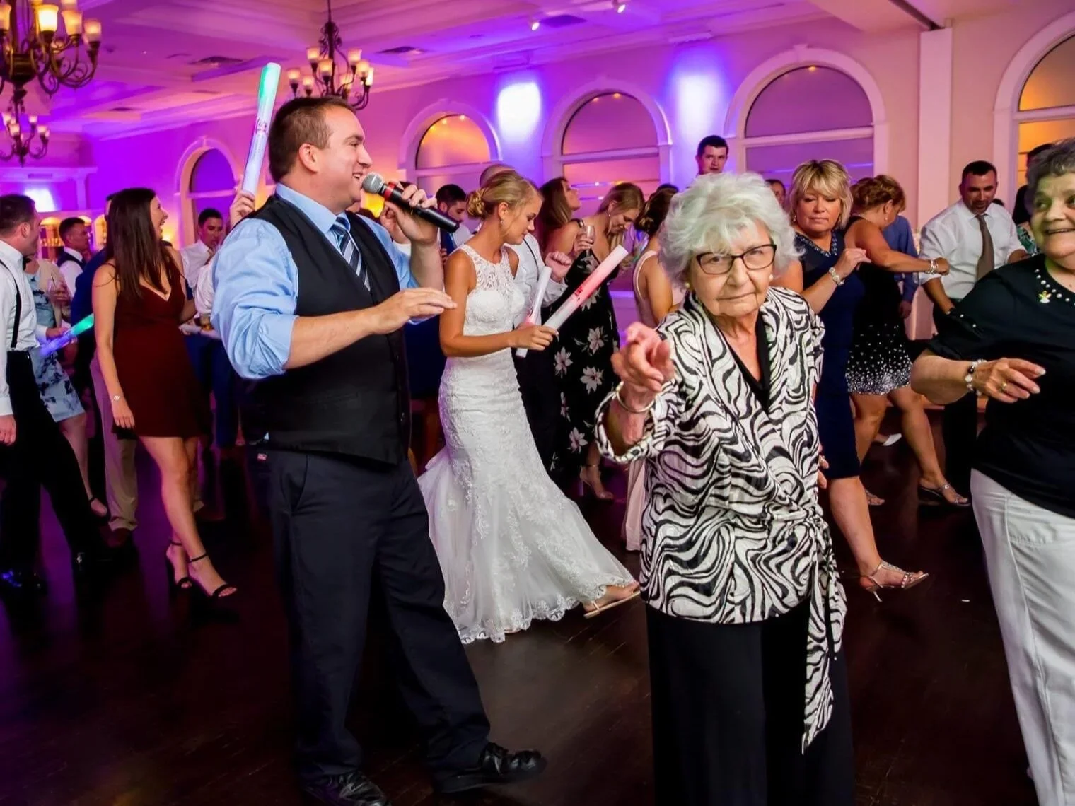 People dancing at a party with purple lighting, some holding glow sticks, including a woman in a white dress and an elderly woman in a zebra print blouse, in a decorated event hall.