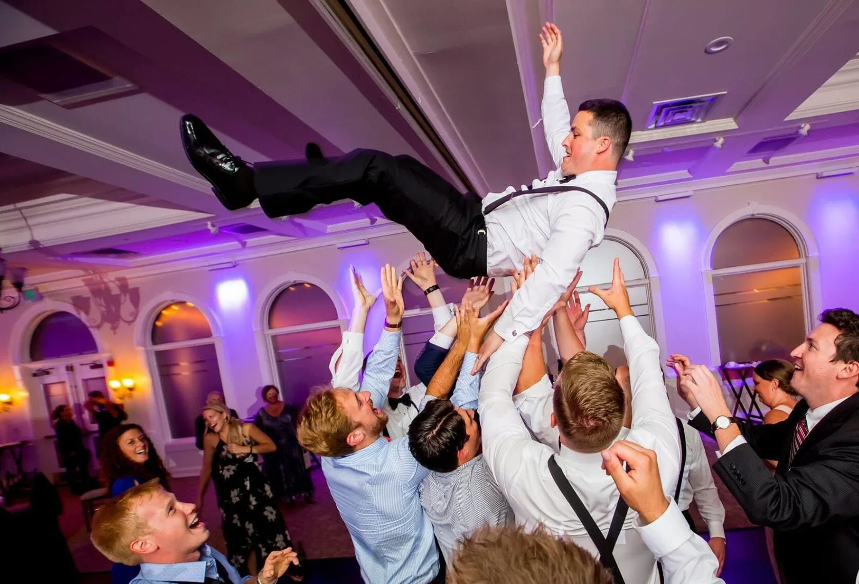 People celebrating at a wedding or party, lifting a man in a white shirt and black suspenders into the air in a decorated banquet hall.