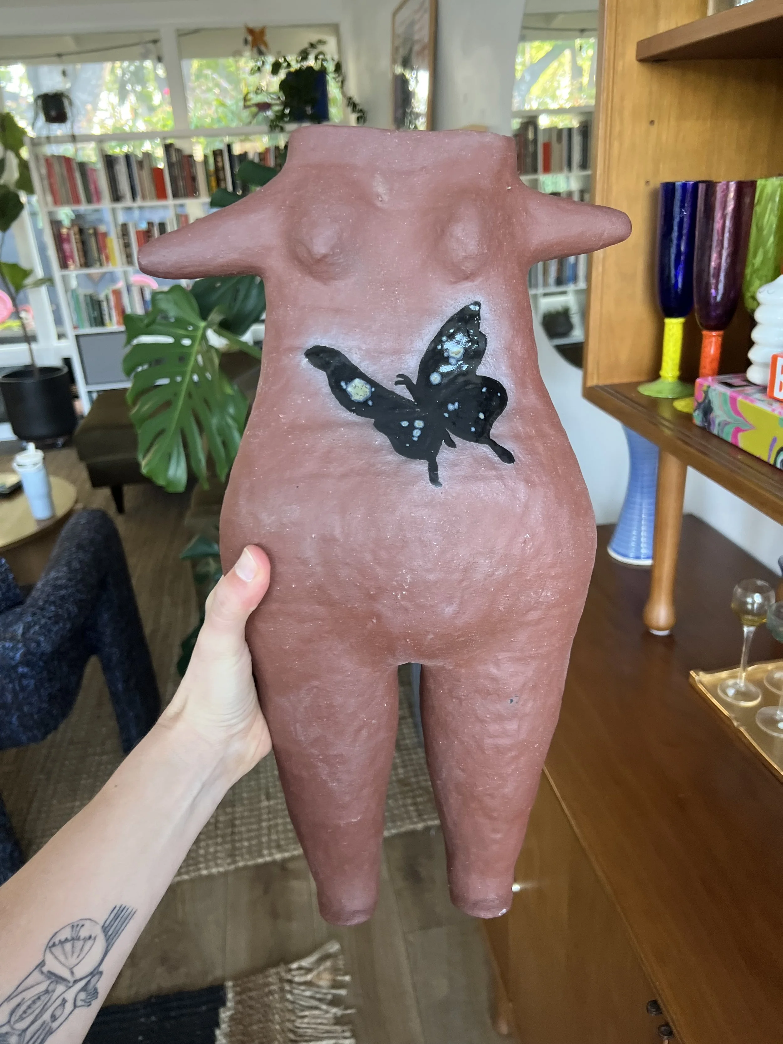 A person holding a terracotta sculpture of a humanoid figure with no head, featuring a black butterfly painted on the chest. The sculpture has a rustic, unglazed finish and is displayed indoors with bookshelves, plants, and colorful glasses in the background.