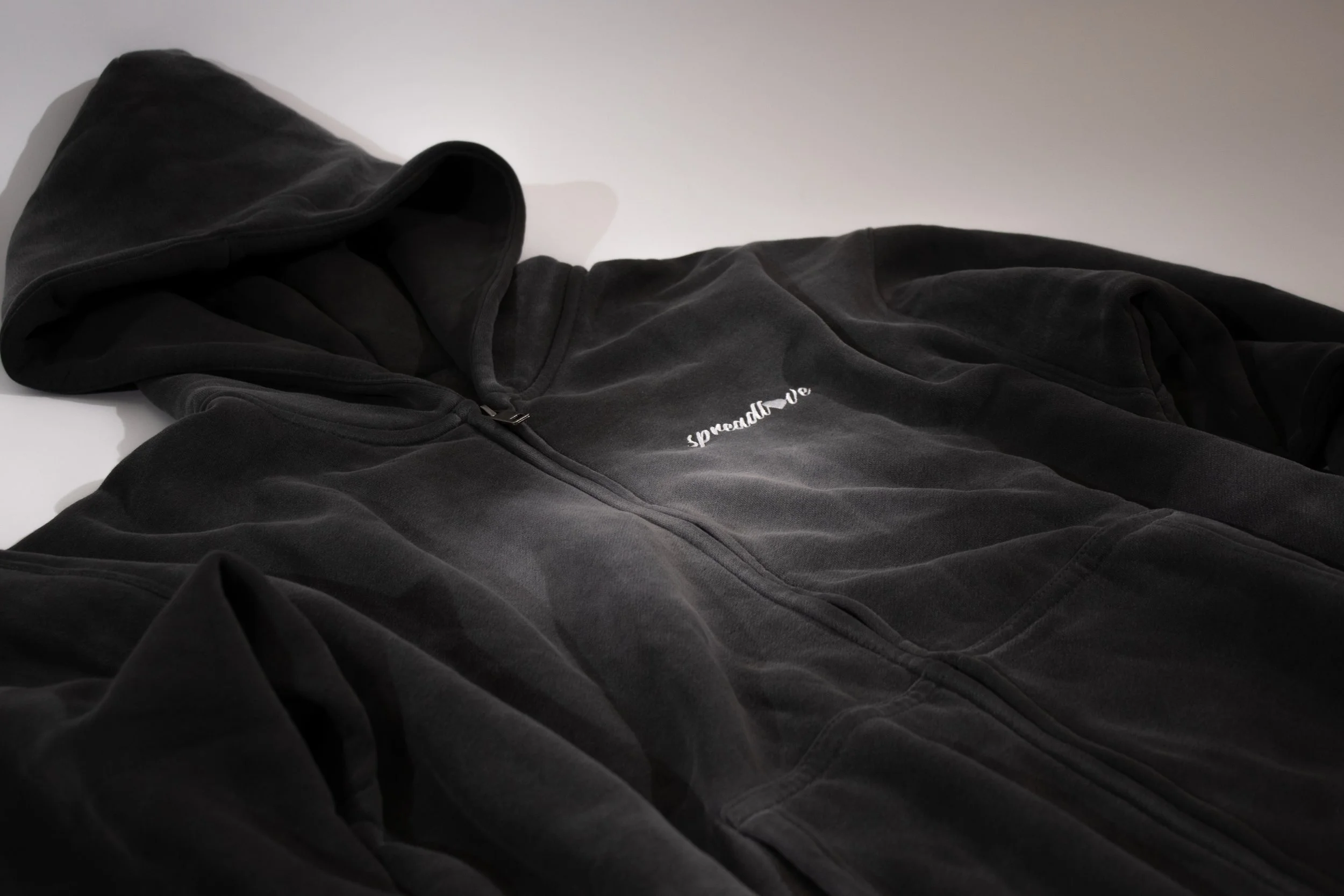 Veste à capuche noire en velours, allongée sur une surface blanche, avec une fermeture éclair au centre et une inscription blanche « speed and love » sur le côté gauche du torse.