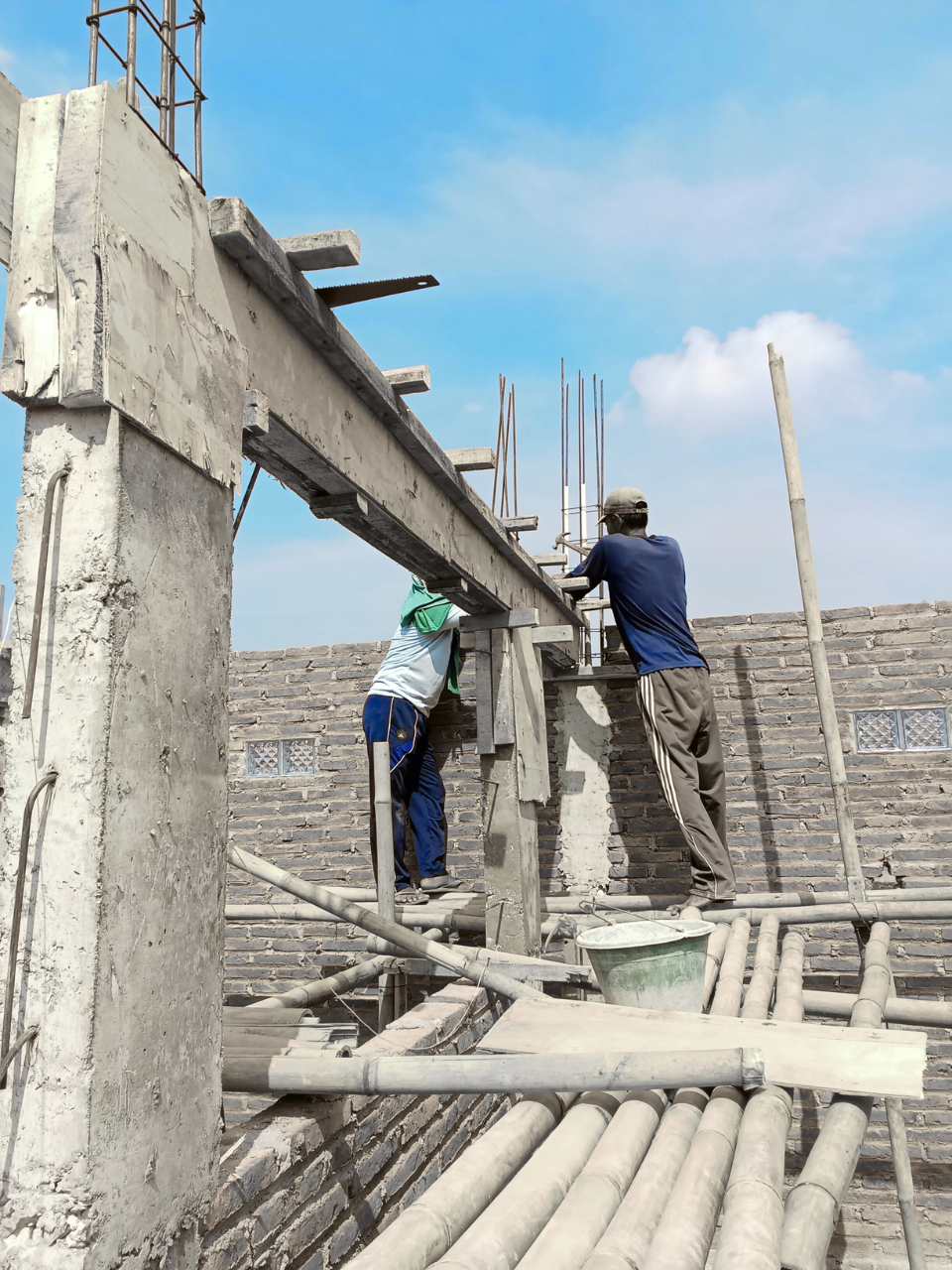 Two construction workers are building a concrete wall on a scaffold at a construction site with a clear blue sky.