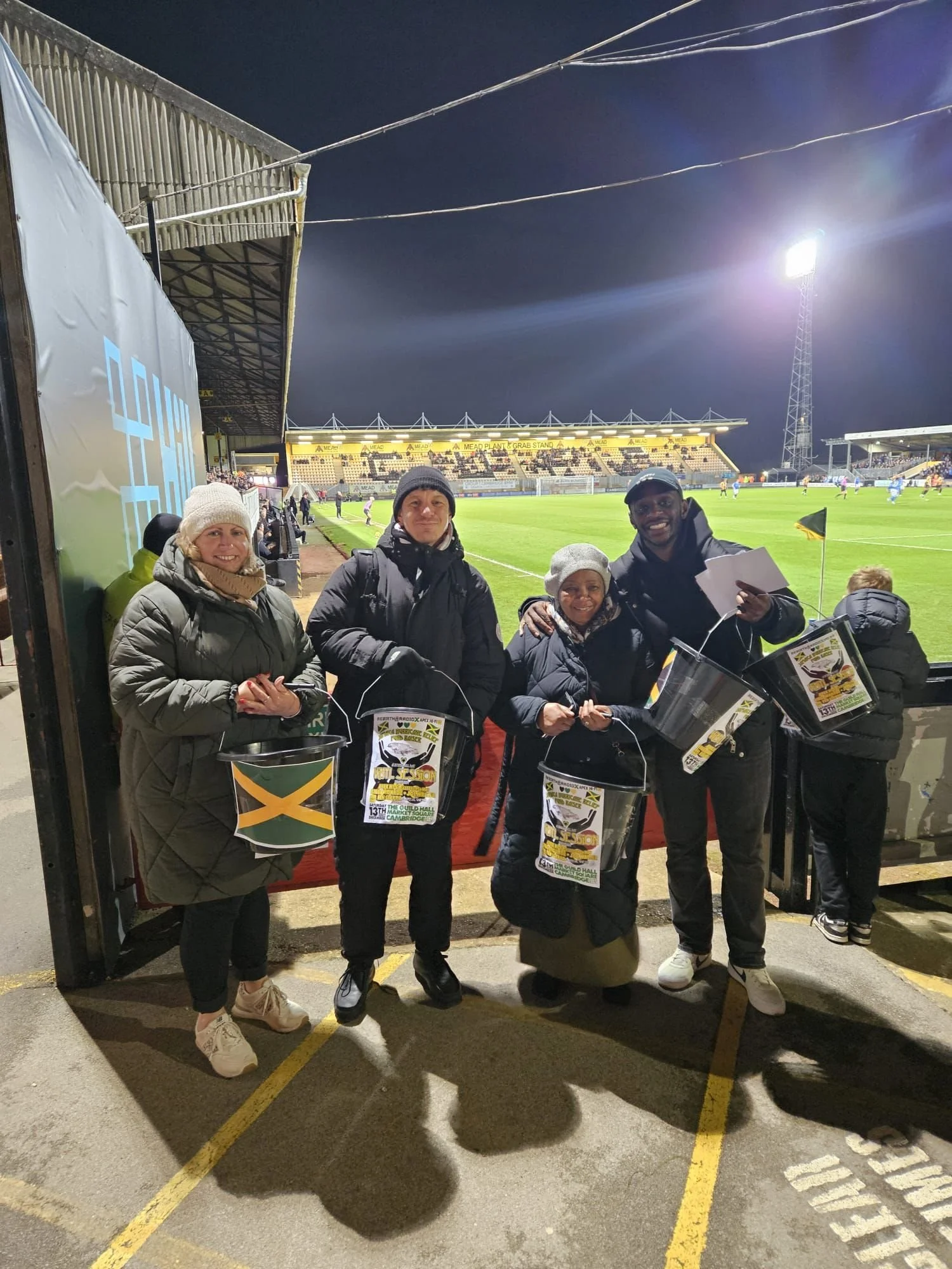 Volunteers fundraising at a football match in Cambridge, holding donation buckets to support Jamaica relief efforts following Hurricane Melissa