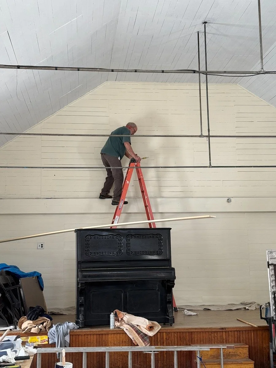 A person standing on a red ladder painting a high wall in a room with a silver ceiling. The room has a black piano on a wooden stage and various items and tools around.