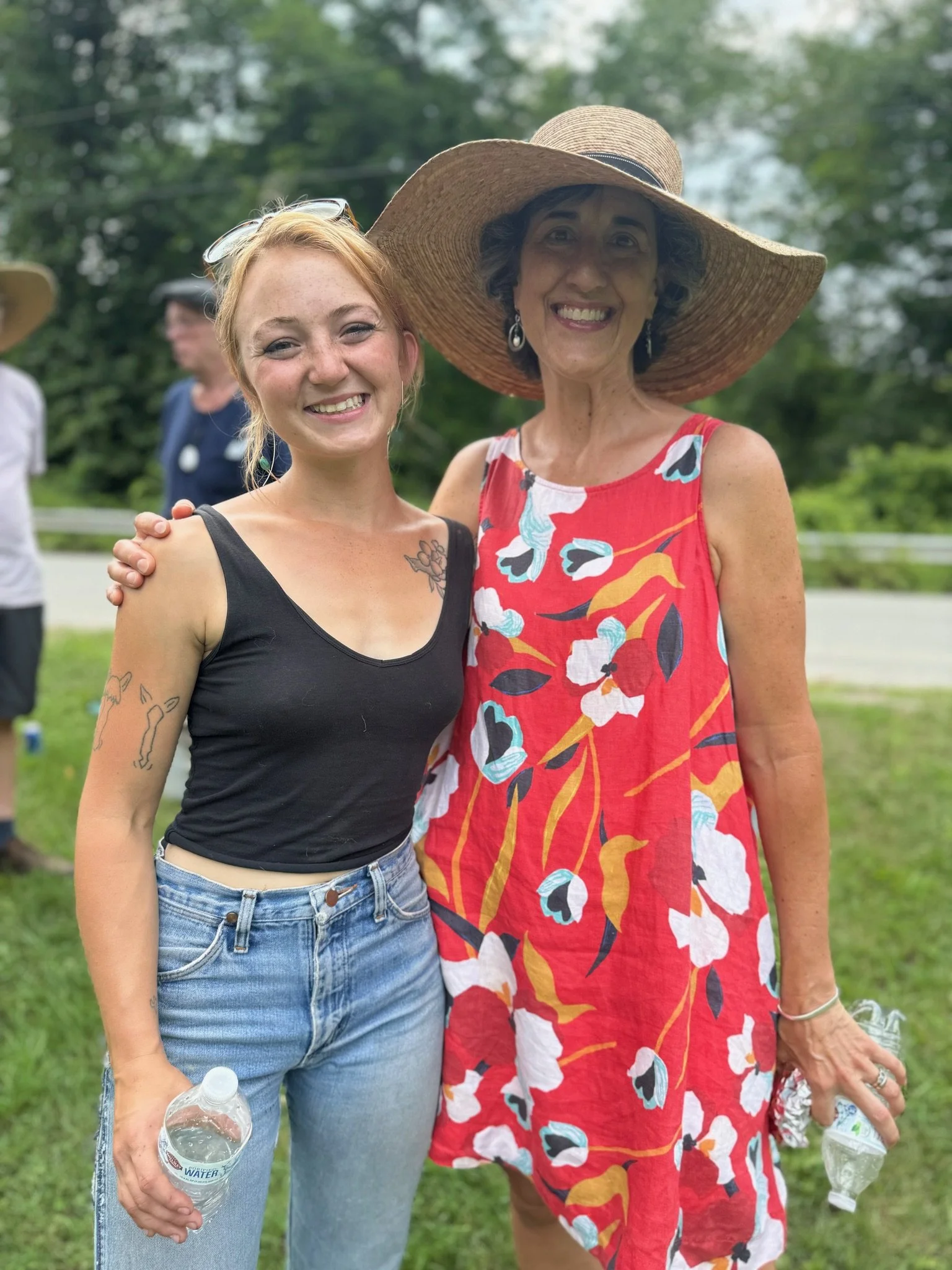 Two women standing outdoors in a park with trees in the background. One woman has curly dark hair, is wearing a large sun hat and a colorful floral dress. The other woman has blond hair, freckles, and is wearing a black tank top and blue jeans, holdi