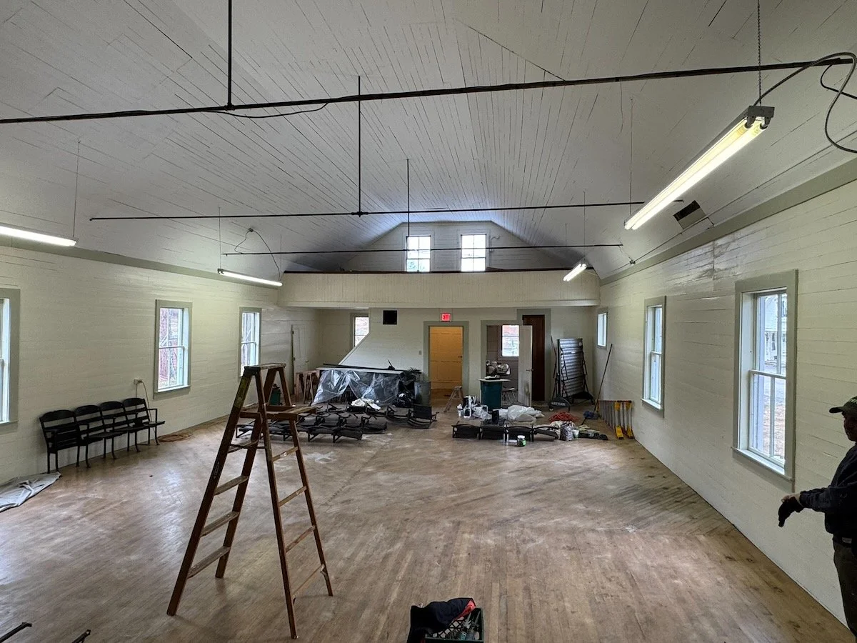 An empty room under renovation with wooden floors, white walls, and several windows. Construction tools, chairs, and a ladder are scattered around.
