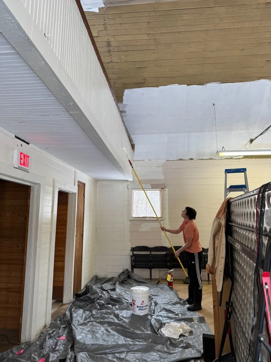 Person painting the ceiling white in a room with plastic covering on the floor, a ladder, and various furniture and equipment.