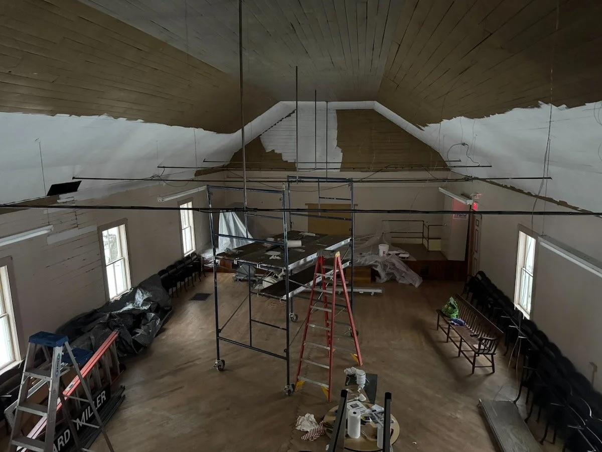 Interior of a room under renovation with dismantled ceiling, scaffolding, ladders, chairs lined up against the walls, and construction materials scattered around.