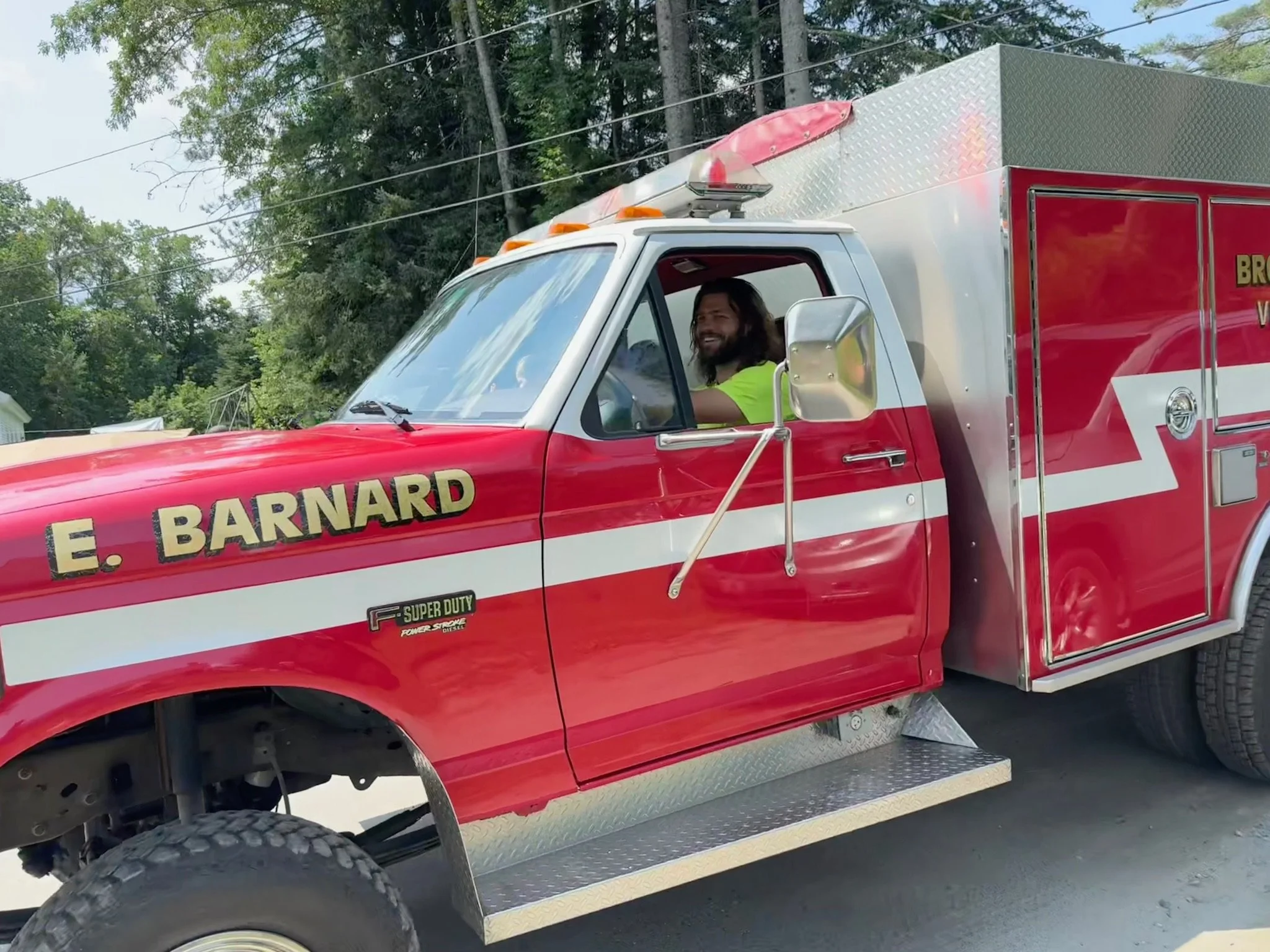Close-up of a red fire truck with a firefighter inside, smiling, surrounded by trees and power lines.